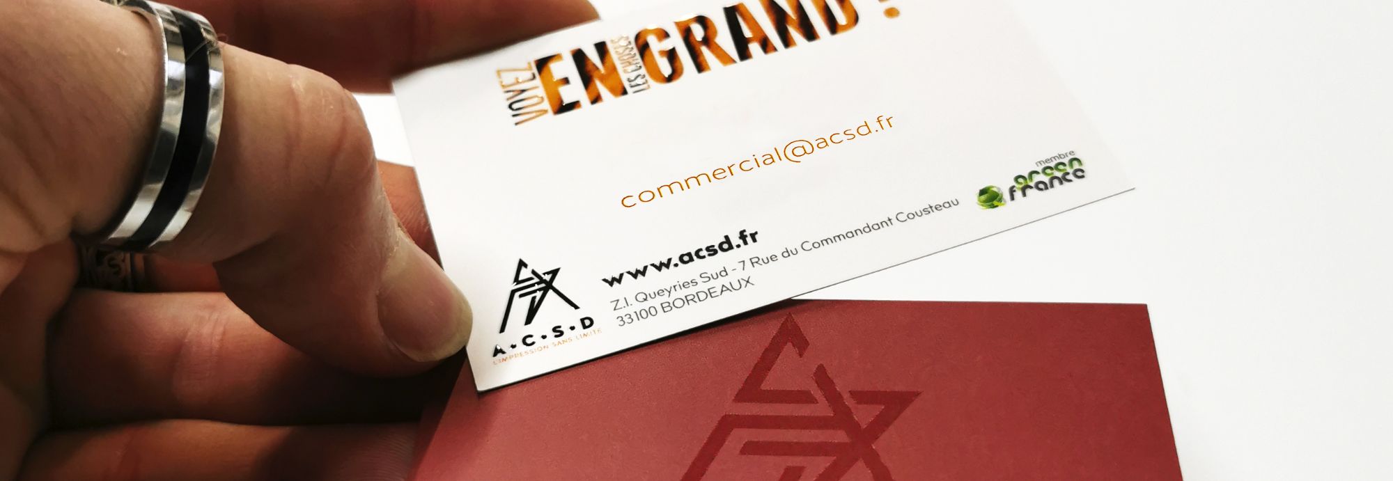 gros plan sur une main tenant la carte de visite de l'imprimerie numérique ACSD pour le service commercial. LOGO ACSD et LOGO green france image-texte en lettres tigrées du slogan voyez les choses en grand